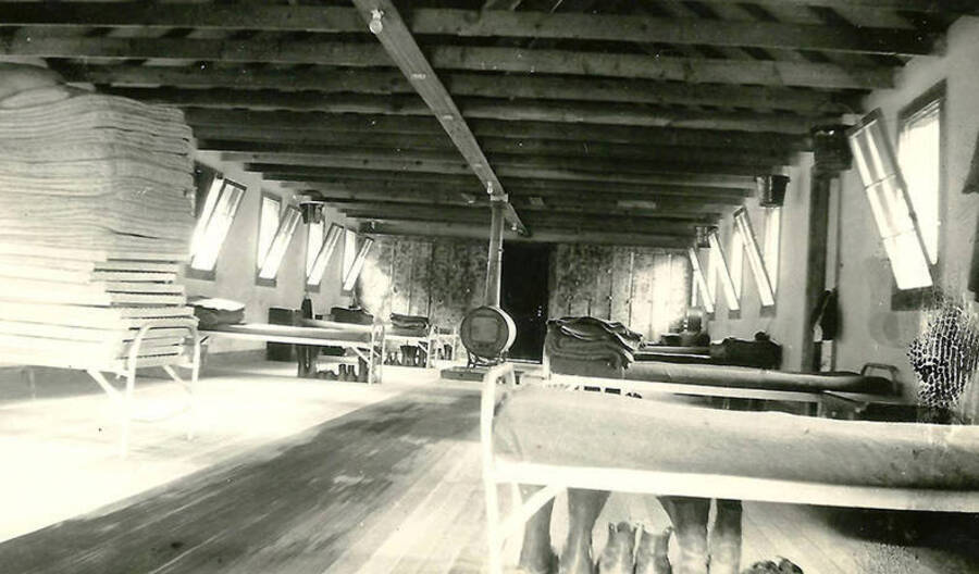An interior view of one of the barracks at CCC Camp Big Creek #2, F-132. The cots are made and extra shoes and boots are laid out in rows underneath them. One of the cots in the foreground has a stack of extra mattresses stacked all the way to the ceiling. The wood stove can also be seen in the middle of the room.