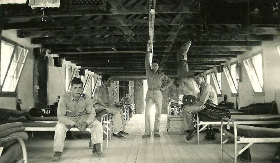 An interior view of one of the barracks at CCC Camp Big Creek #2, F-132. The men are lounging about on the cots and one is hanging upside-down from the ceiling from a wooden beam. One has a pipe in-between his teeth. Back of the photo reads: 'Barracks all cleaned up for inspection.'
