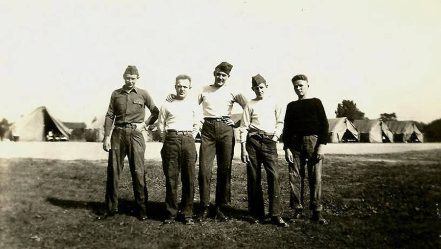 Five CCC men standing outside a row of tent barracks. Back of the photo reads: 'Tent camp while barracks were being built'.