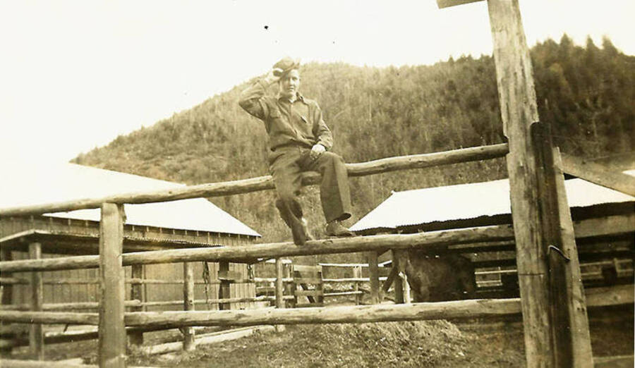 A CCC man tips his hat at the camera and perches atop the fence of the corral at CCC Camp Big Creek #2, F-132. A horse can be seen through the rungs. Back of the photo reads: 'I'm an old cowhand! Corral at Co. 531'