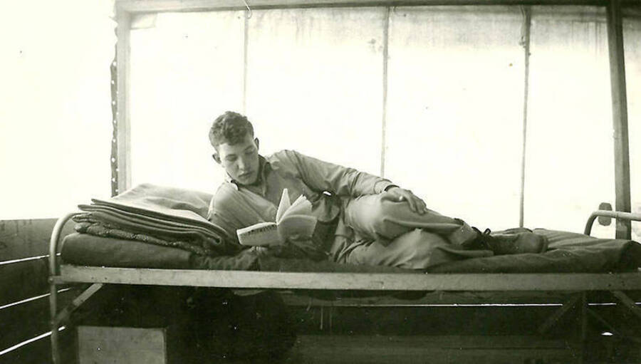 A CCC man lounges on his cot, reading a book, inside of a tent barrack at CCC  Spike Camp near Mullan, Idaho. Back of the photo reads: 'After a hard day's work at Spike Camp Mullan, Ida.'