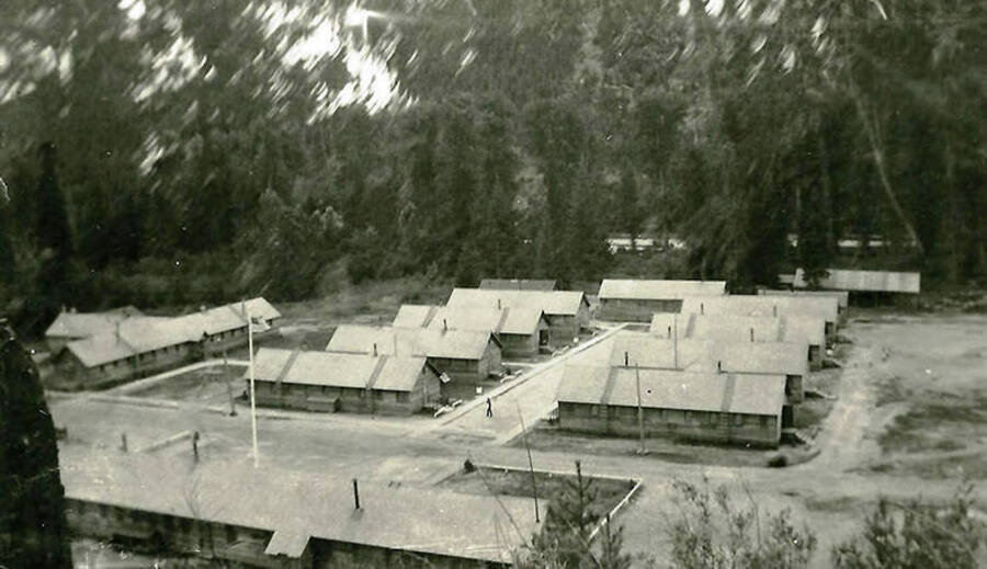 A view of CCC Camp Big Creek #2, F-132. Headquarters building sits in the foreground while the barracks are in two rows in the middle of the photo. Back of the photo reads: 'Looking down on Company area CCC Co. 531. Coeur d'Alene River in the background.'