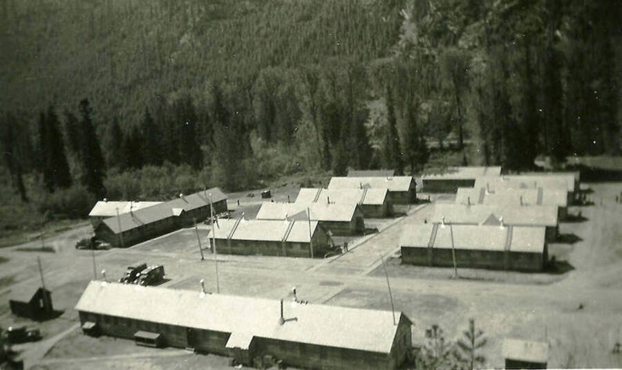 A view of CCC Camp Big Creek #2, F-132. Headquarters building sits in the foreground while the barracks are in two rows in the middle of the photo. Back of the photo reads: 'Rec Hall in foreground. Barracks T-shaped building to left is messhall'.
