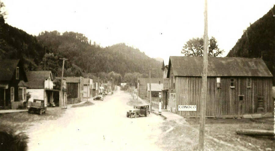A view of a mining town nearby CCC Camp Big Creek #2, F-132. A sign on one of the buildings reads: 'Conoco' and there are a couple automobiles parked on the side of the road. Back of the photo reads: 'Lead and silver mining town 1938/39'.
