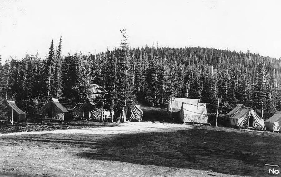 A view of the tent barracks and field at Camp Black Bear. Taken in August. 'Port of Bertha Hill in background. First four tents on left are officers quarters. Next 3 tents are first, Educational Advisor's tent, First Aid tent, and Forestry personnel tent. Near in is Library tent and Truck supply tent.' Description of photos says Camp number is P-262.