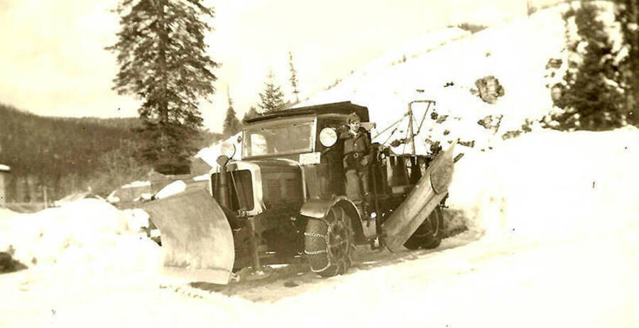 A CCC man poses on the side of a snow plow near CCC Camp Big Creek #2, F-132. The plow  is attached to a large truck which has chains on its tires. Back of the photo reads: 'Ready for winter?'