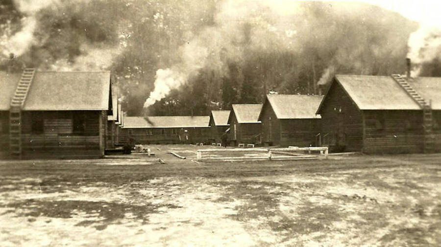 A view of CCC Camp Big Creek #2, F-132. Headquarters building is in the background while the barracks are in two rows in the middle of the photo. Smoke is rising from many of the chimneys. Back of the photo reads: 'Cold weather all the stoves are working.'