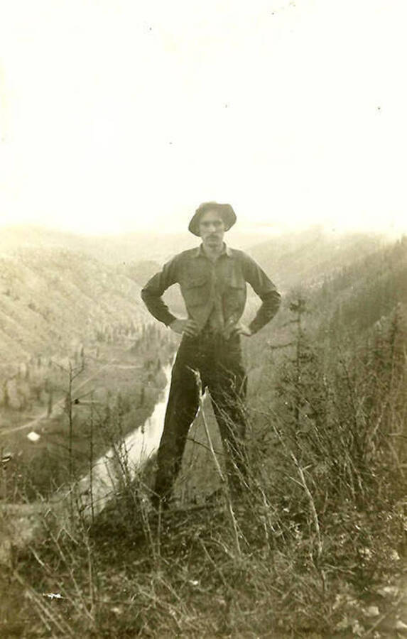 A CCC man stands above the valley and the North Fork of the Coeur d'Alene River and CCC Camp, F-132. Back of the photo reads: 'On the edge. The river is 'way' down below. Some mountain'.