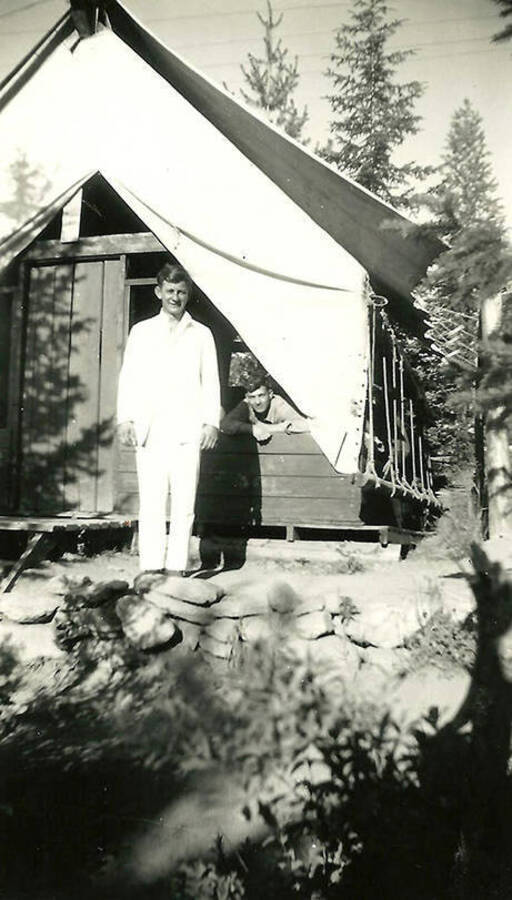Two CCC men inside and in front of a tent barrack at the Spike Camp in Mullan, Idaho. The man standing in front of the tent is wearing a cook's outfit. Back of the photo reads: 'One of our cooks at Spike Camp - Mullan.'
