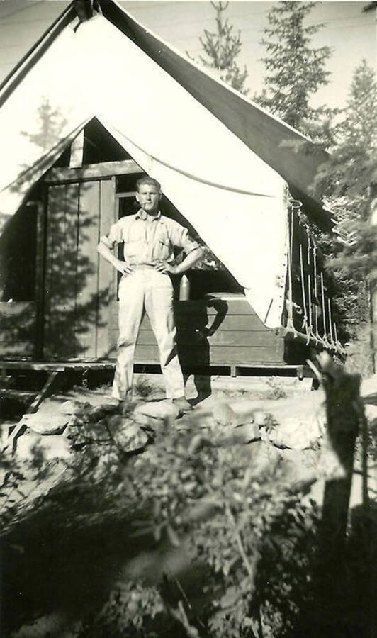 A CCC man stands in front of a tent barrack at the spike camp in Mullan, Idaho. Back of the photo reads: 'One of our leaders at Mullan spike camp. We built Shoshone Co. Park near there. Esther and I visited the park in 1978. It was still in good shape.'