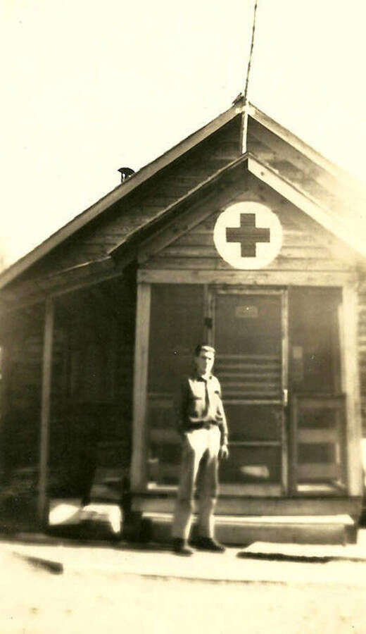 A CCC man stands outside a building at CCC Camp Big Creek #2, F-132, with the sign of the red cross above the door.