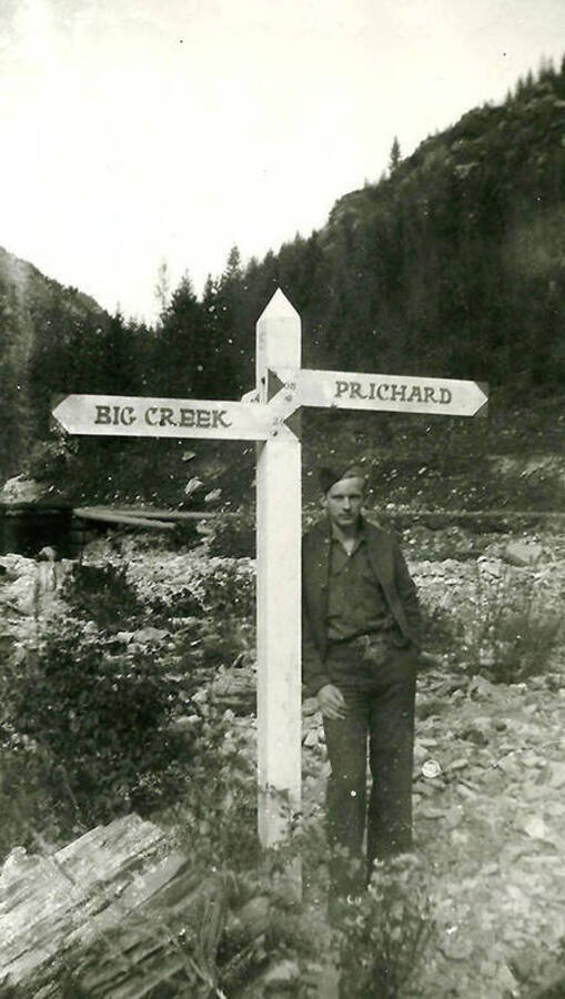A CCC man poses next to a road sign in front of a creek and tree-covered hill. The sign reads: 'Big Creek' on the left and 'Prichard' on the right. Back of the photo reads: 'We didn't need a sign to tell us where we were, 'nowhere'.'