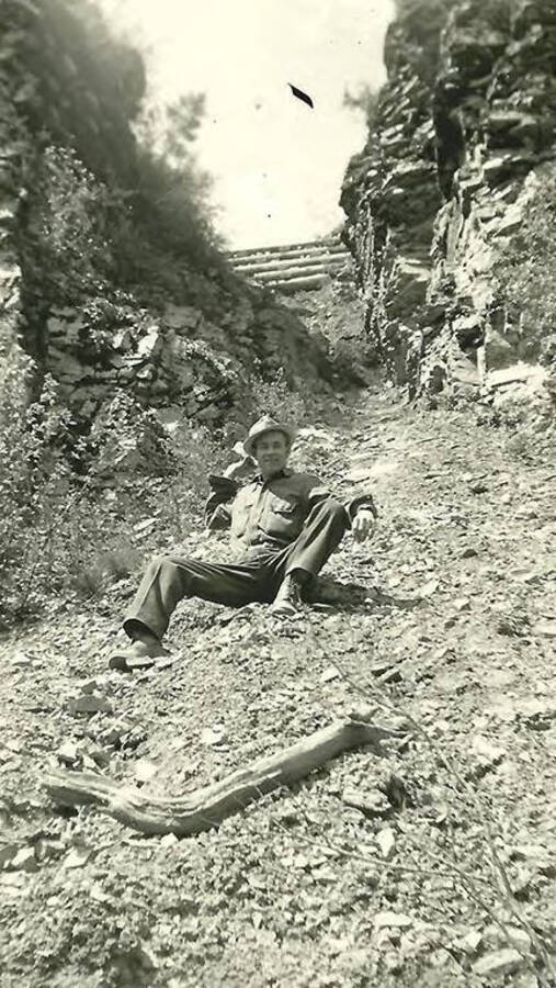 A CCC man poses on a steep slope, a rocky gulley in between two cliff faces. The top of the gulley has logs laid horizontally across it. The man has a rock in his hand, cocked back, ready to throw. Back of the photo reads: 'Rough country (Leader) Bill Whitley from Kentucky'