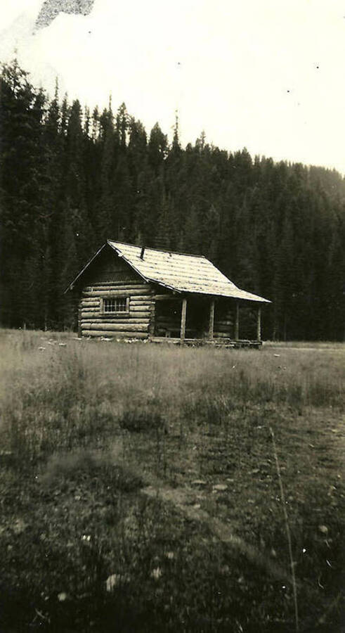 A log cabin stands in a meadow in front of a wall of trees near CCC Camp Big Creek #2, F-132. Back of the photo reads: 'Probably the Forest Ranger's cabin. He was near camp.'