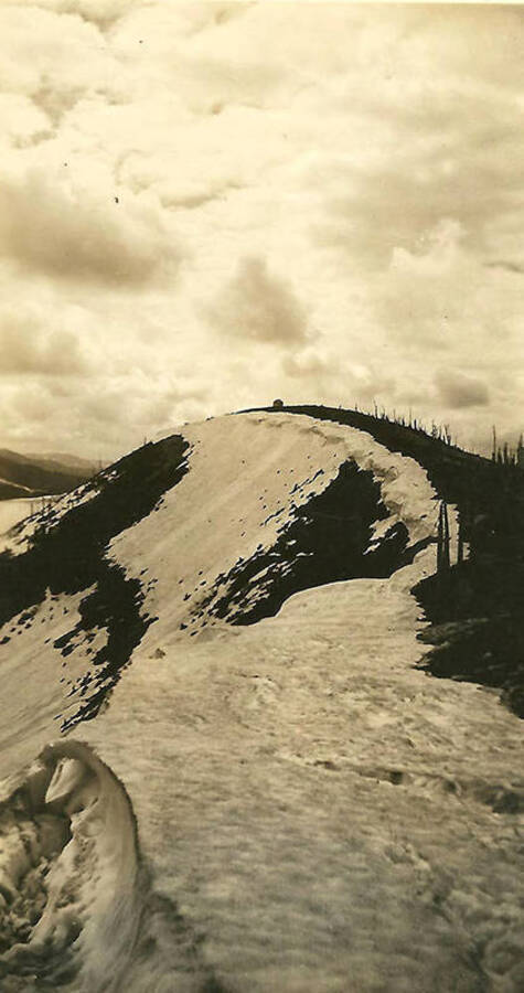 A view of a snow-covered hill. There appears to be a small building on the top of the hill. Back of the photo reads: 'near Sunset Peak, looking South toward Kellogg.'