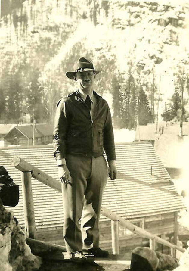 A CCC man stands on the side of a hill at the same height as the CCC Camp behind him the camp is Big Creek #2, F-132. Back of the photo reads: 'One of our 'foremen' at CCC Co. 531'.