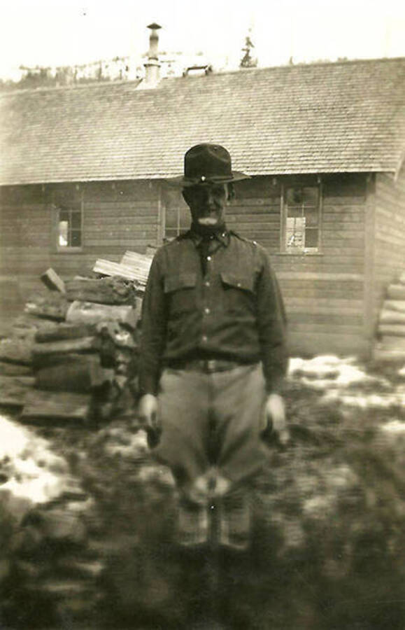 A CCC officer stands in front of a building and firewood pile at CCC Camp Big Creek #2, F-132. Back of the photo reads: 'St. Salu one of our Co. 531 officers'.