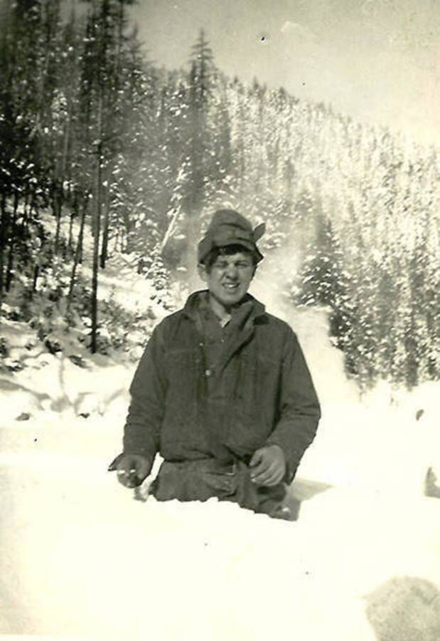 A CCC man poses in the snow that comes up to his hips near CCC Camp Big Creek #2, F-132. A snow-covered forest rises on a hill in the background. Back of the photo reads: 'Joe Bugaj?? 3ft of snow near main camp Big Creek'.
