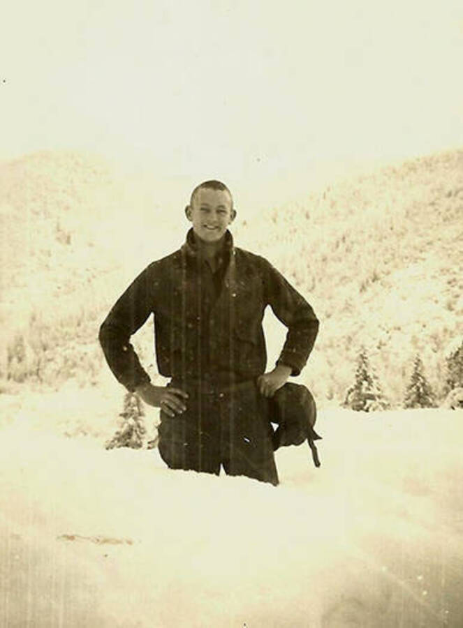 A CCC man poses in the snow that comes up to his thighs near CCC Camp Big Creek #2, F-132. A snow-covered forest rises on a hill in the background. Back of the photo reads: 'I think the snow is deep enough!'
