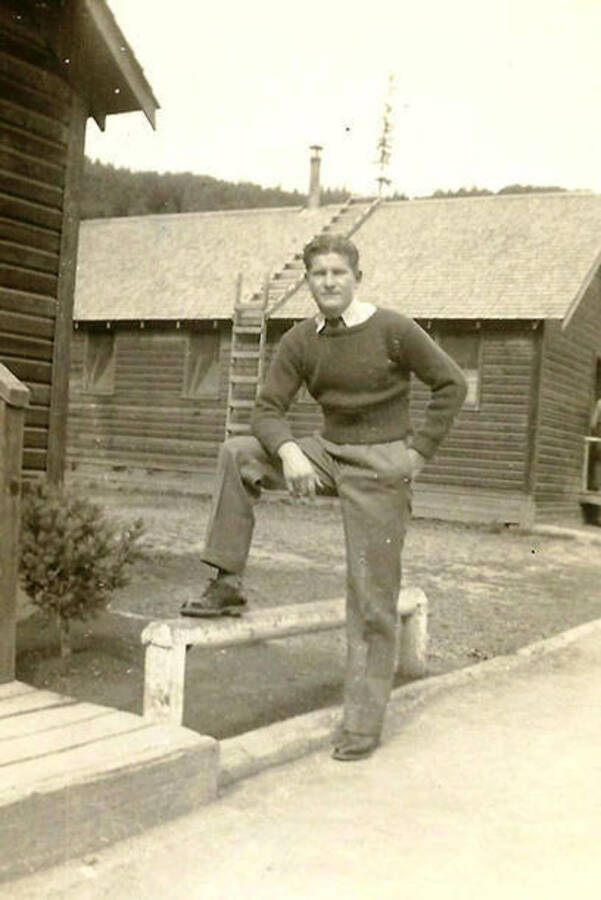 A CCC man poses with his leg up on a ledge next to the barracks in CCC Camp Big Creek #2, F-132. The barrack behind him has a ladder that leads up the side of the building and onto the roof.
