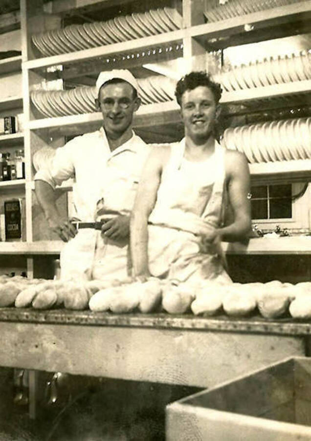 Two CCC men standing in the kitchen in front of rows of plates and behind a counter full of peeled potatoes. Back of the photo reads: 'man on right [undecipherable word] Johnny Muscar camp Baker. Can't remember his ass't. Met Johnny in Portland, Me 1943 He was in U.S.C.G.'