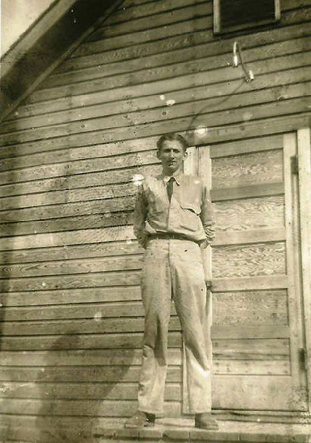 A CCC man stands in front of the barracks at CCC Camp Big Creek Camp #2, F-132. Back of the photo reads: ''Red' Emry'