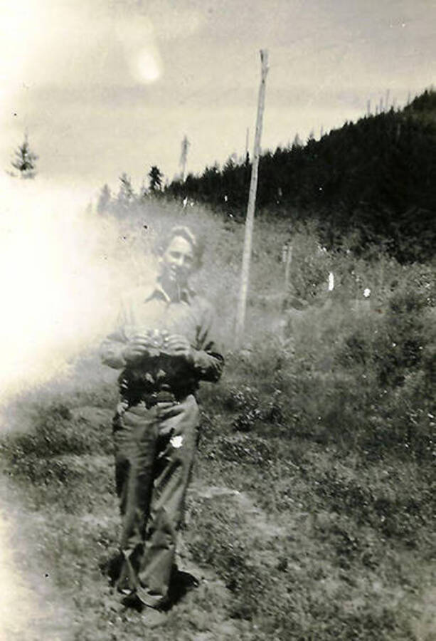 A CCC man standing in the woods under a telephone pole with a camera in his hand. The camera is pointed outward, as if the photographers were taking portraits of each other.