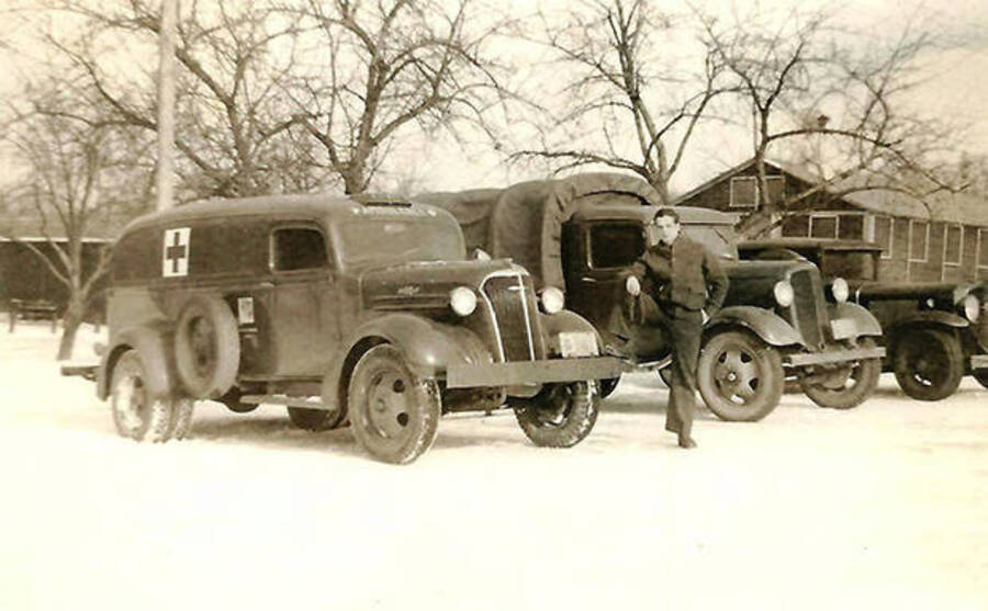 A CCC man in front of several trucks, including an ambulance, at CCC Camp Big Creek #2, F-132. A building can be seen in the background. Snow covers the ground. Back of the photo reads: 'Great old trucks mostly 37 Chev.'