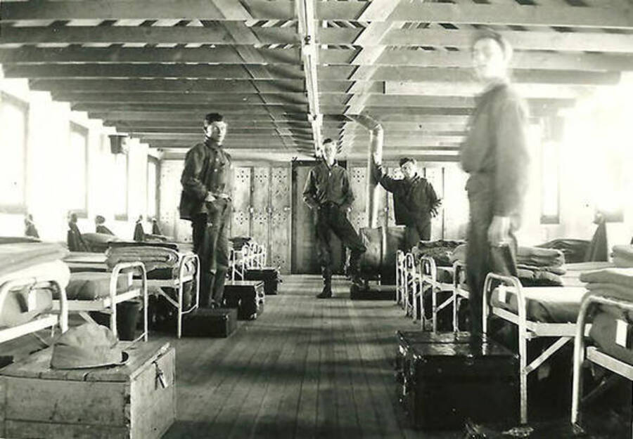 Four CCC men lounging in the barracks at CCC Camp Big Creek Camp #2, F-132. The barrack is full of cots, chests, lockers and extra blankets. Back of the photo reads: 'Our home away from home. Note the bare rafters. Nothing fancy here'.