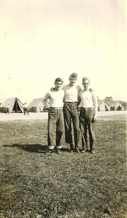 Three CCC men pose in front of tent barracks with several other men in the background.