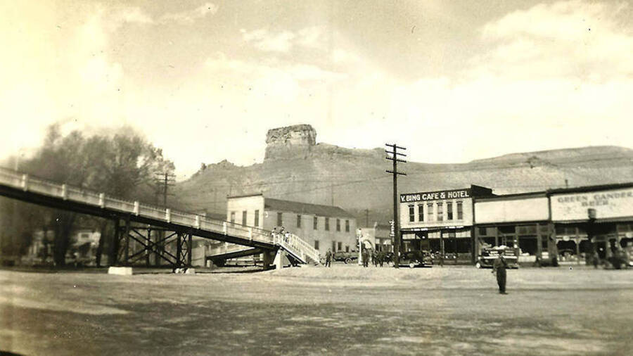 CCC men stand around in a town square with a towering rock face in the background. A pedestrian bridge spans the road. Two storefronts read as follows: 'Y. Bing Cafe & Hotel' and 'Green Gander Beer'