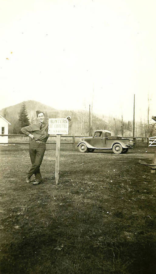 A CCC man leans against a sign in front of a truck in a field at CCC Camp Big Creek #2, F-132. The sign reads: 'Hunters Be Careful CCC Men Working'.