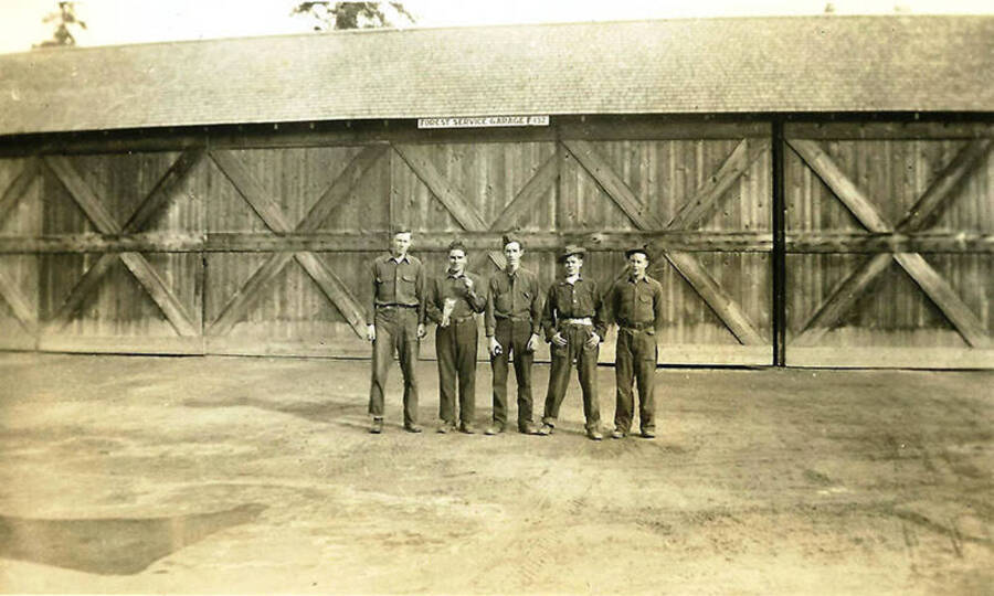 Five CCC men standing outside in front of CCC Big Creek Camp #2, F-132 Garage. There are large sliding doors behind them and a sign that reads: 'Forest Service Garage F-132'
