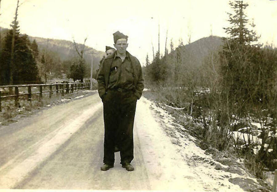 Two CCC men standing on a snowy road with two forested hills in the background. One enrollee is peeking out behind the shoulder of the other.