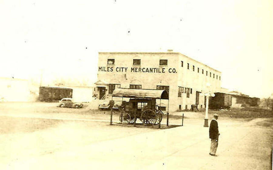 A man stands out in front of the 'Miles City Mercantile Co.' There is also a sheltered carriage, a truck, and a couple of train cargo cars.
