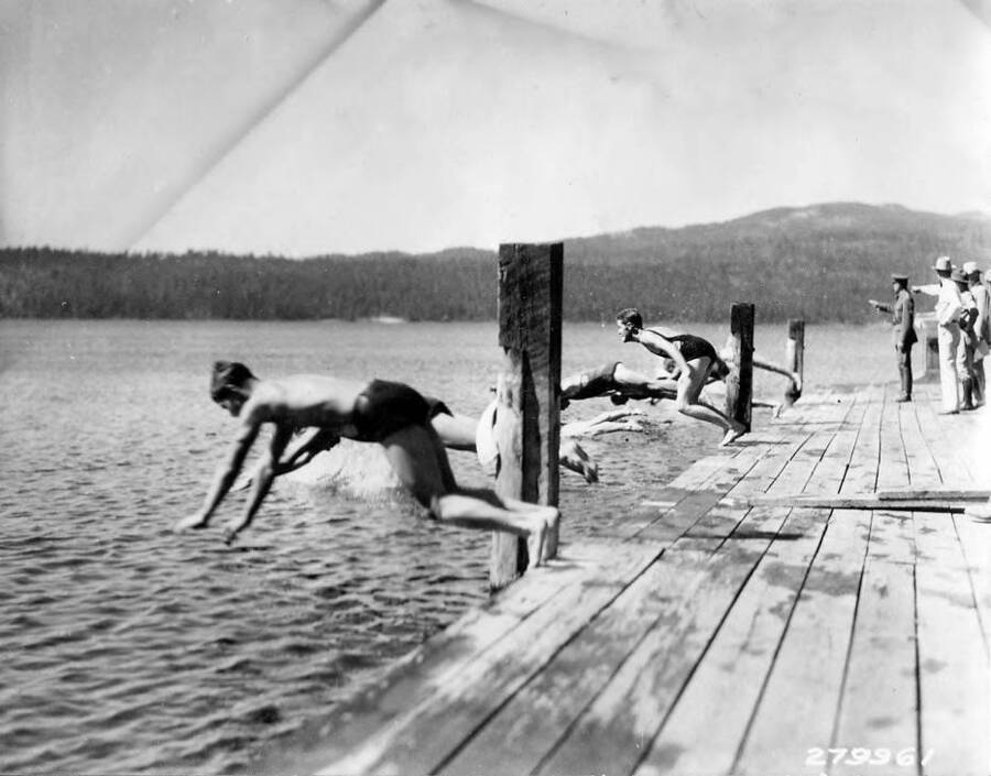 Young men dive into the lake from the pier during the swimming race at the CCC Carnival in McCall, Idaho. Observers in the background point to something across the lake.