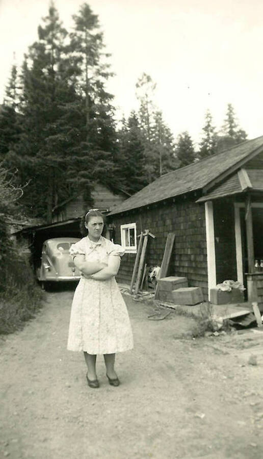 A woman stands in a driveway with her arms crossed. A house and car are in the background. Back of the photo reads: 'Fairlynn Foster, Mullan, Idaho 1939. Father Louie Foster and I were good friends.'