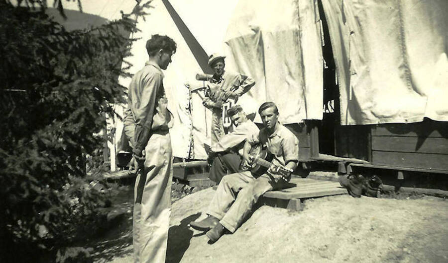 Four CCC men lounge outside of a tent barrack. One is holding a guitar. A sign outside the barrack reads: 'A'. Back of the photo reads: 'Elya Burg (Staffy) playing guitar in spike camp.