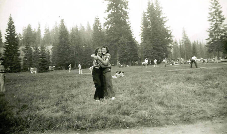 Two young women hug each other in the outfield of a CCC Baseball Game. Back of the photo reads: 'Just a couple of the girls who came to the park from Burke Canyon (a small town near Mullan).