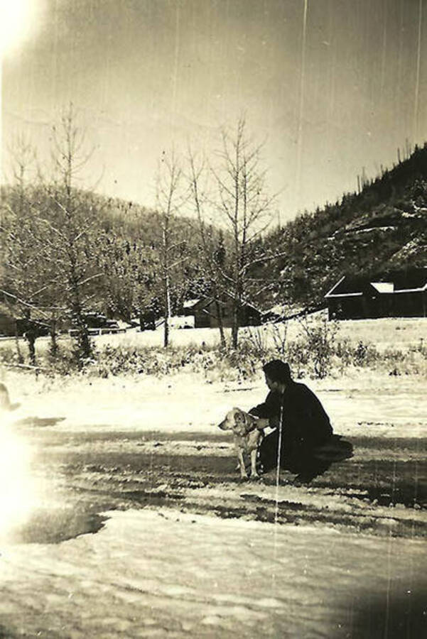 A CCC man poses on a snowy road with his dog in front of CCC Camp Big Creek #2, F-132. The camp buildings and forested hills can be seen in the background. Back of the photo reads: 'All camps had a dog'.