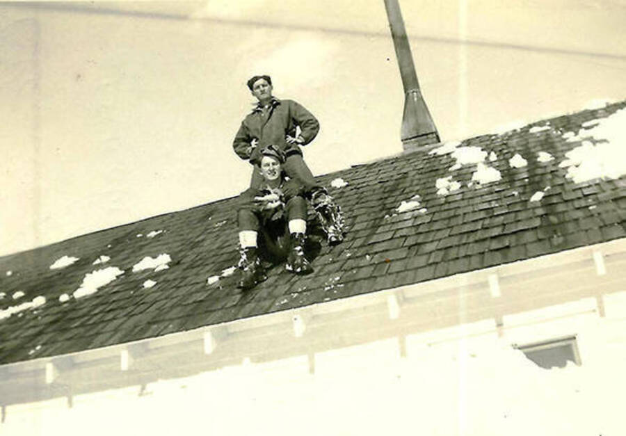 Two CCC men perch atop a snow-covered roof at CCC Camp Big Creek Camp #2, F-132. Back of the photo reads: 'Cleaning off the barracks roof - you can just see the corner of the window to the right - some snow!'