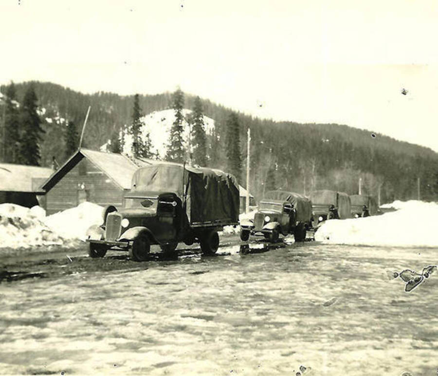 Four covered trucks driving into a snow covered CCC Camp, Big Creek #2, F-132. Back of the photo reads: 'On the way to a big weekend in Wallace, Idaho'.