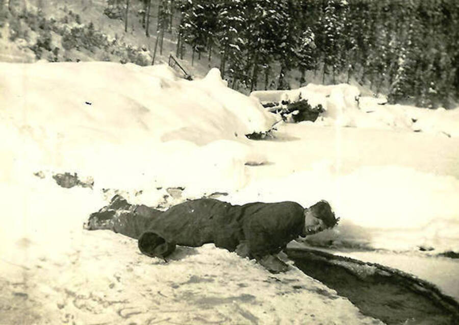 A CCC man lies on his stomach in the snow near a hole in the ice. Back of the photo reads: 'Boy am I thirsty!'