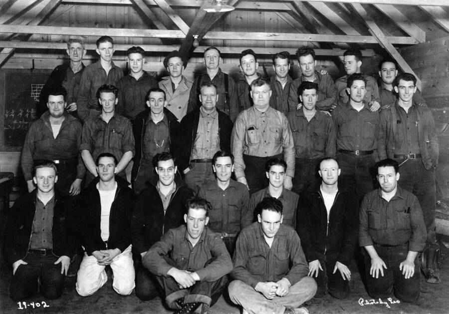 Group portrait of Camp Troy personnel, Company 244, Troy, Idaho. Writing on photo reads '19-402 Photo by Leo' There are also fractions and a musical clef on the blackboard behind the men.