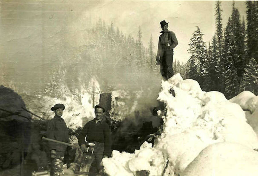 Three CCC men pose at work in the woods. One man carries an ax, and another stands atop a snow pile. Back of the photo reads: 'Very narrow valley. We were cutting out an old log jam that turned the river against our road and was washing it away. Leader Bill Whitley on top of snow pile.'