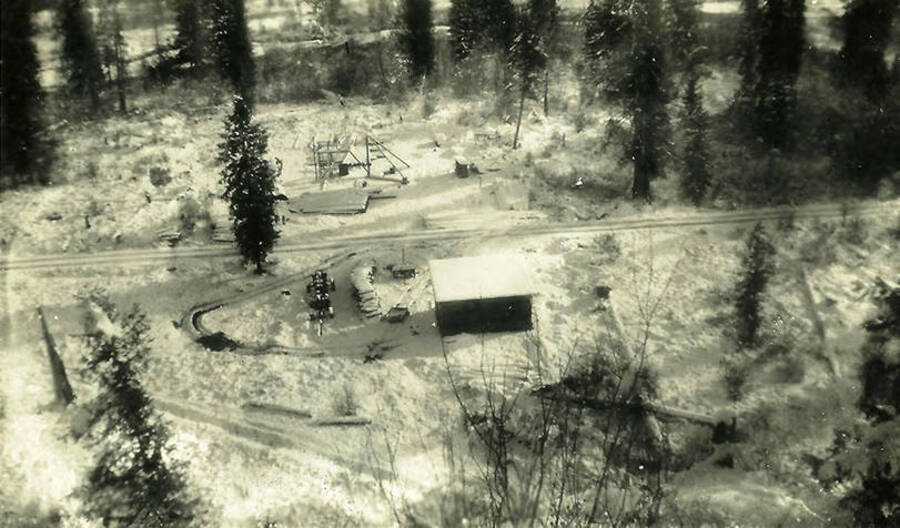 A snow-covered road runs past a building, woodpile and tractor as well as other unidentified structures. Back of the photo reads: 'Our camp area, including maint bldg in foreground. Coeur d'Alene River in background. All grown up now in 50 to 60 yr old pines.
