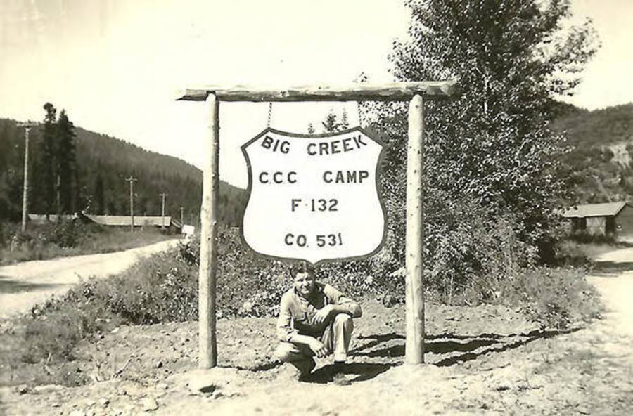 A CCC man poses underneath a sign for CCC Camp Big Creek #2, it reads: 'Big Creek CCC Camp F-132 Co. 531'. The Forest Ranger Garage and the barracks can be seen on either side of the photo in the background. Back of the photo reads: ''Red' (Can't remember name) The F-132 was our forestry camp designation. HQ was at Ft. George Wright in Spokane, Wash.'