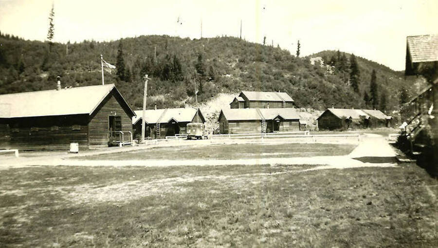 A view of the courtyard of CCC Camp Big Creek #2, F-132. There are several buildings and a flagpole around the edge of the courtyard and a truck parked on the edge. One of the buildings is an infirmary. Hills rise in the background. Back of the photo reads: 'Camp Big Creek'
