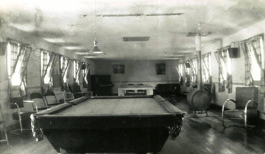 A view of the recreation room at CCC Camp Big Creek #2, F-132. Pool table dominates the foreground with chairs  and a camp stove lining the walls. A piano sits in one corner with a couch in the other. Back of photo reads: 'Our 'rec' hall. You had a choice ping-pong, pool, or just B.S. each other. We went to the movies in Wallace; once a month.'