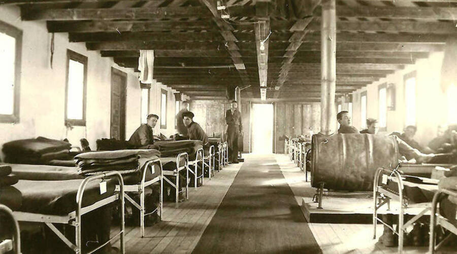 Six CCC men sitting in the barracks at CCC Camp Big Creek #2, F-132, on their cots. Extra blankets are stacked on the beds and a camp stove in the center of the room. Back of the photo reads: 'More barracks. You can see our 'heating' system. We cut and split every stick of wood we burned; both for heat and the cookin' in the kitchen too. It took a lot.'